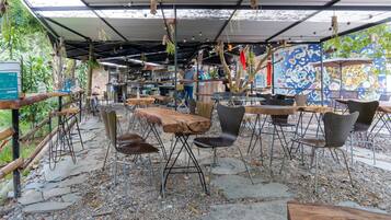 Un café al aire libre con mesas de madera rústica y sillas de metal, rodeado de un sendero de grava y vegetación.