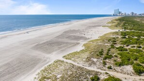 Een kuststrand met een zandstrand, schaarse begroeiing en een verafgelegen stadsachtergrond.