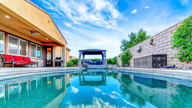 A swimming pool with a covered area, a barbecue grill, and red chairs.