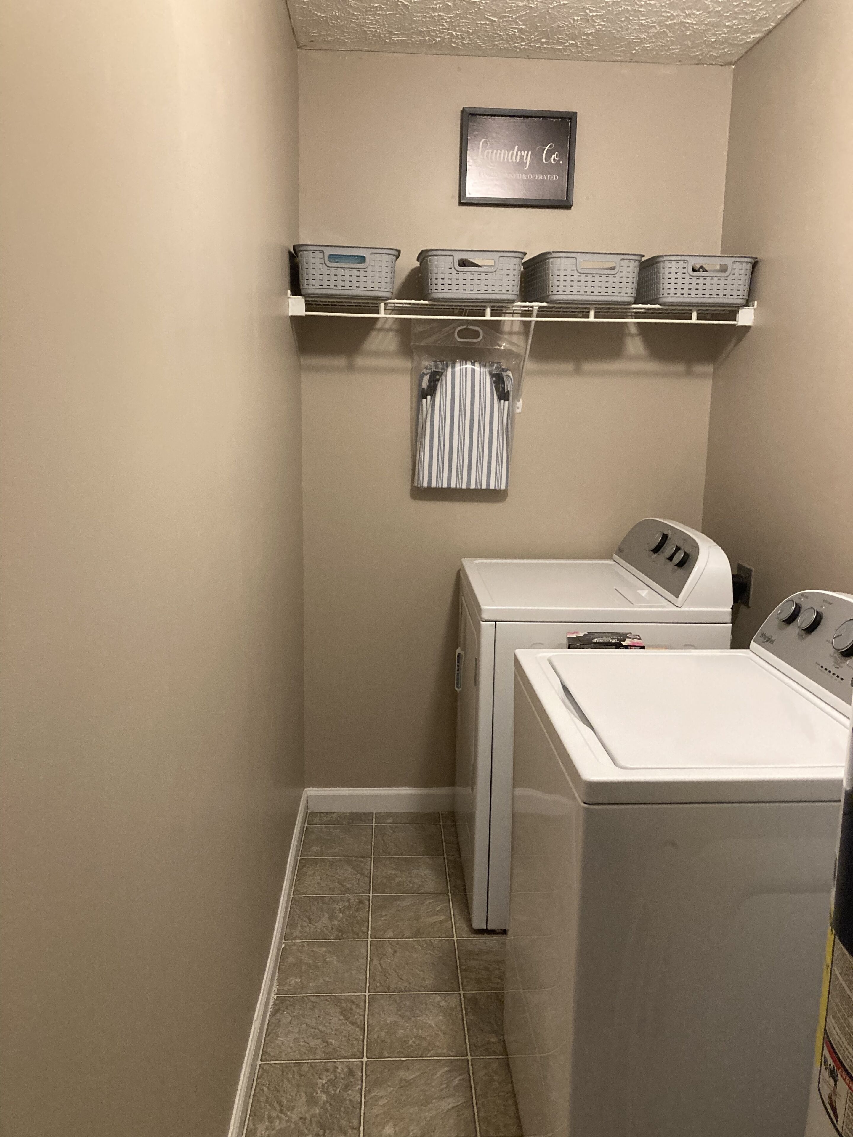 A laundry room with two front-loading washing machines, a shelf with laundry baskets, and a wall-mounted sign.
