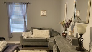 A living room with a grey sofa, white fluffy pillows, a wooden coffee table, a white shag rug, a wooden dresser, a mirror, and a vase with flowers.