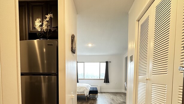 A hallway with a wooden floor, a stainless steel refrigerator, a bed with a white comforter, and a window with curtains. - Sophisticated Studio Apartment in Gadsden, AL (Gadsden)