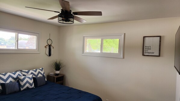 A bedroom with a bed, two windows, a ceiling fan, a wall-mounted plant, and a motivational quote poster.