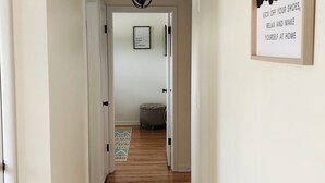 A hallway with wooden flooring, white walls, and a ceiling light fixture. A framed welcome sign is visible on the right wall. - Cozy Comfy (Bourbonnais)
