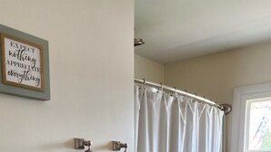 A bathroom with a shower curtain, a towel rack with two towels, a framed inspirational quote, and a window with a view of greenery. - Cozy Comfy (Bourbonnais)