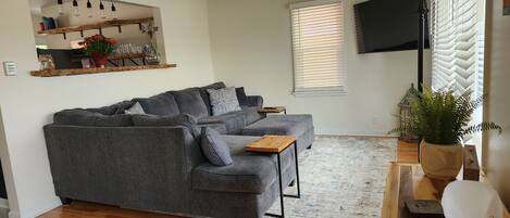 A modern living room with a grey sectional sofa, a wooden coffee table, and a large flat-screen TV mounted on the wall.