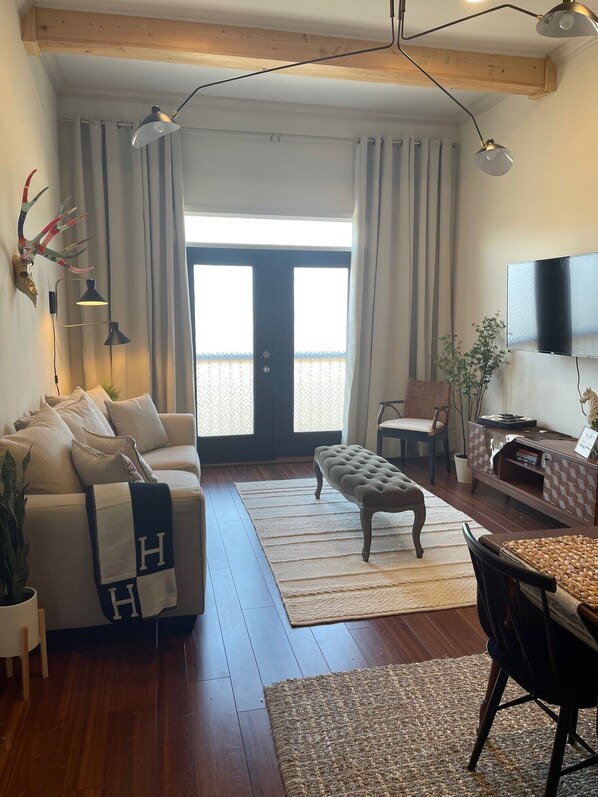 A living room with a sofa, ottoman, dining table, and chairs. There is a television mounted on the wall, a plant, and a wooden ceiling.