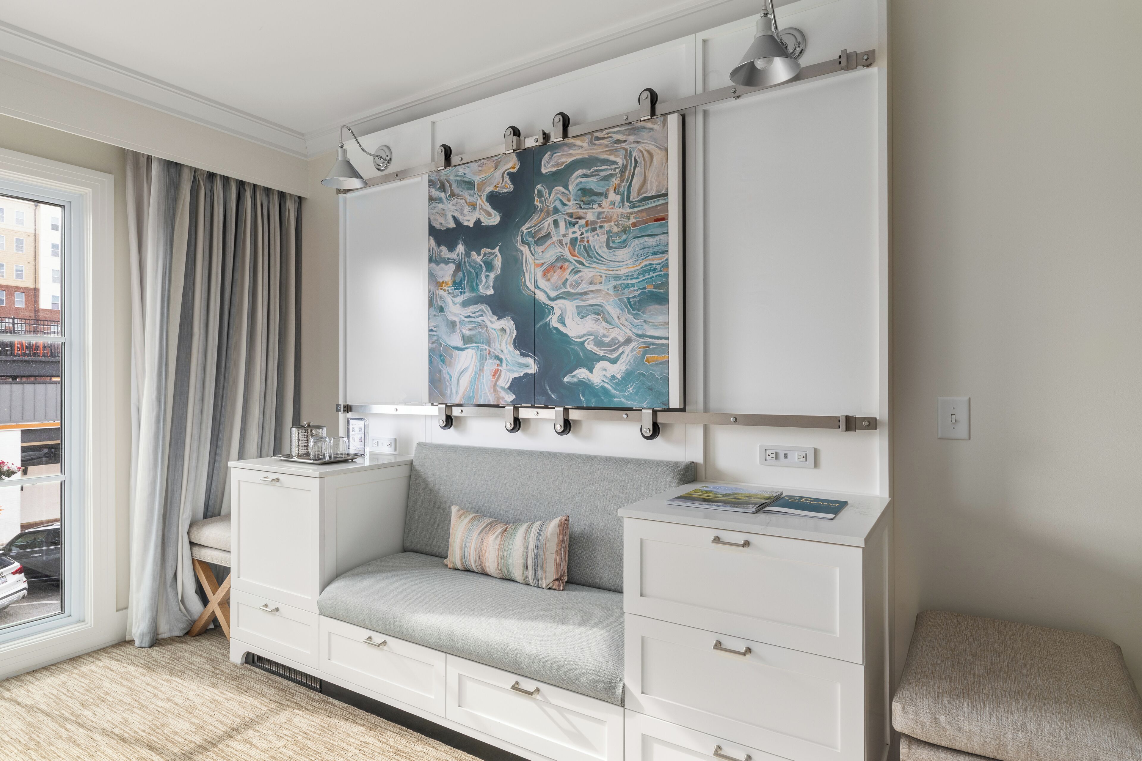 Een moderne hotelkamer met een grijze bank, witte opbergmeubels en een groot abstract schilderij aan de muur.