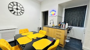A room with a round table, yellow chairs, a black clock, a neon sign, a sink, and cabinets.