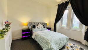 A bedroom with a bed, nightstands, a lamp, a window with curtains, and a plant on a dresser.
