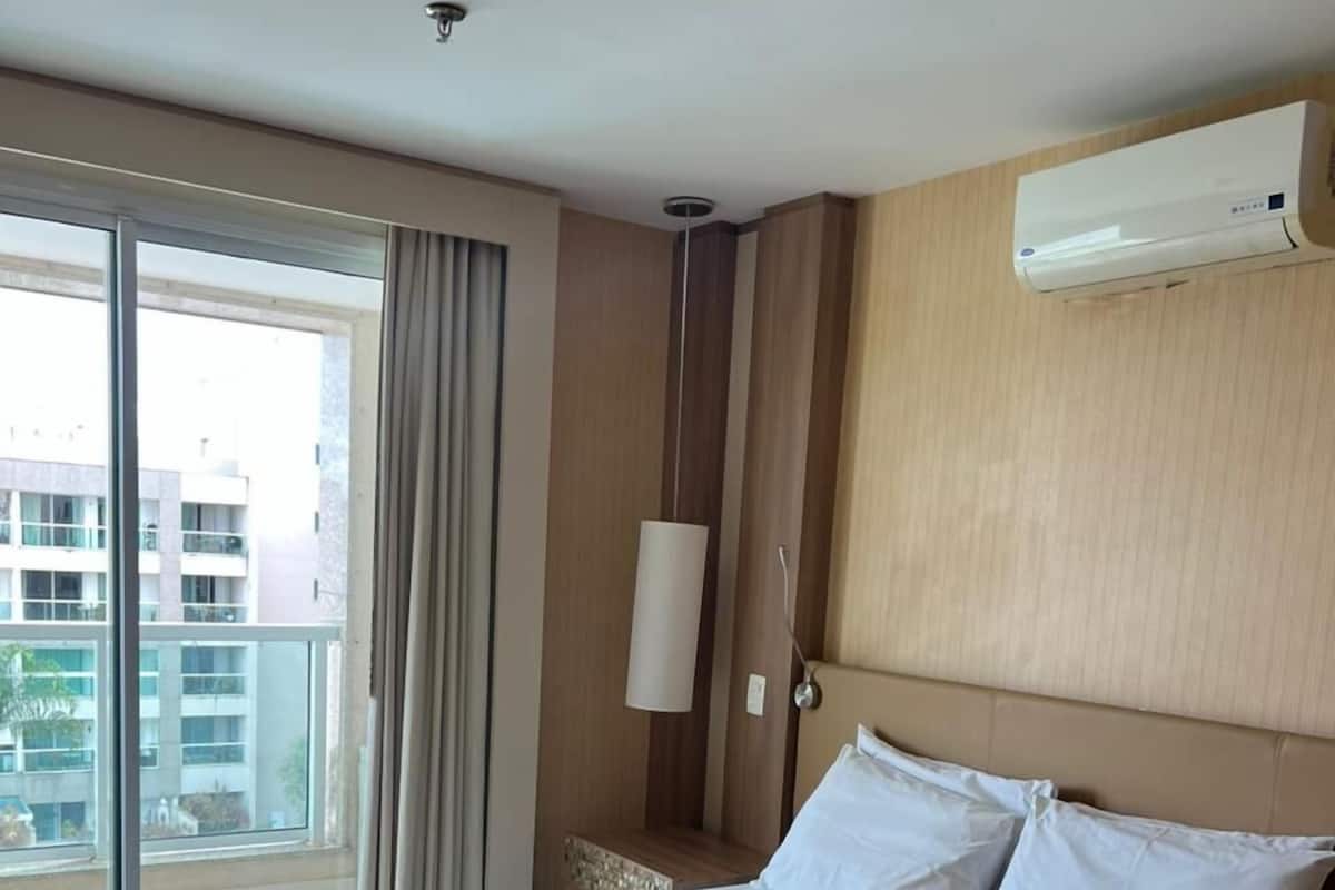 A hotel room with a large bed, a view of the city through the window, and an air conditioning unit on the wall.