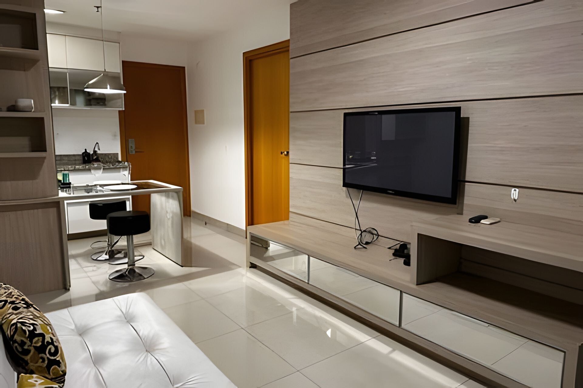 Una sala moderna con un televisor de pantalla plana montado en una pared de madera, una zona de cocina con armarios blancos y un sofá con cojines estampados.