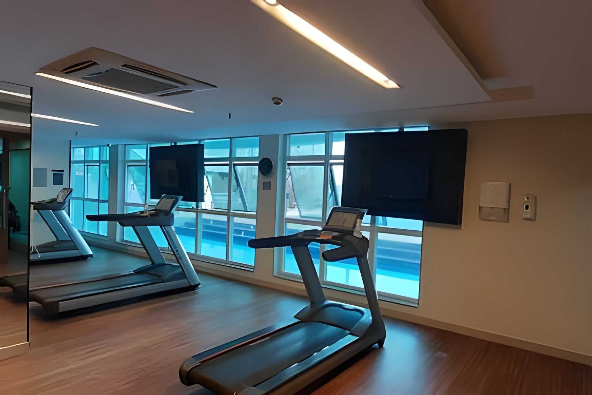 A gym with treadmills and a large mirror, large windows, and a flat-screen TV on the wall.