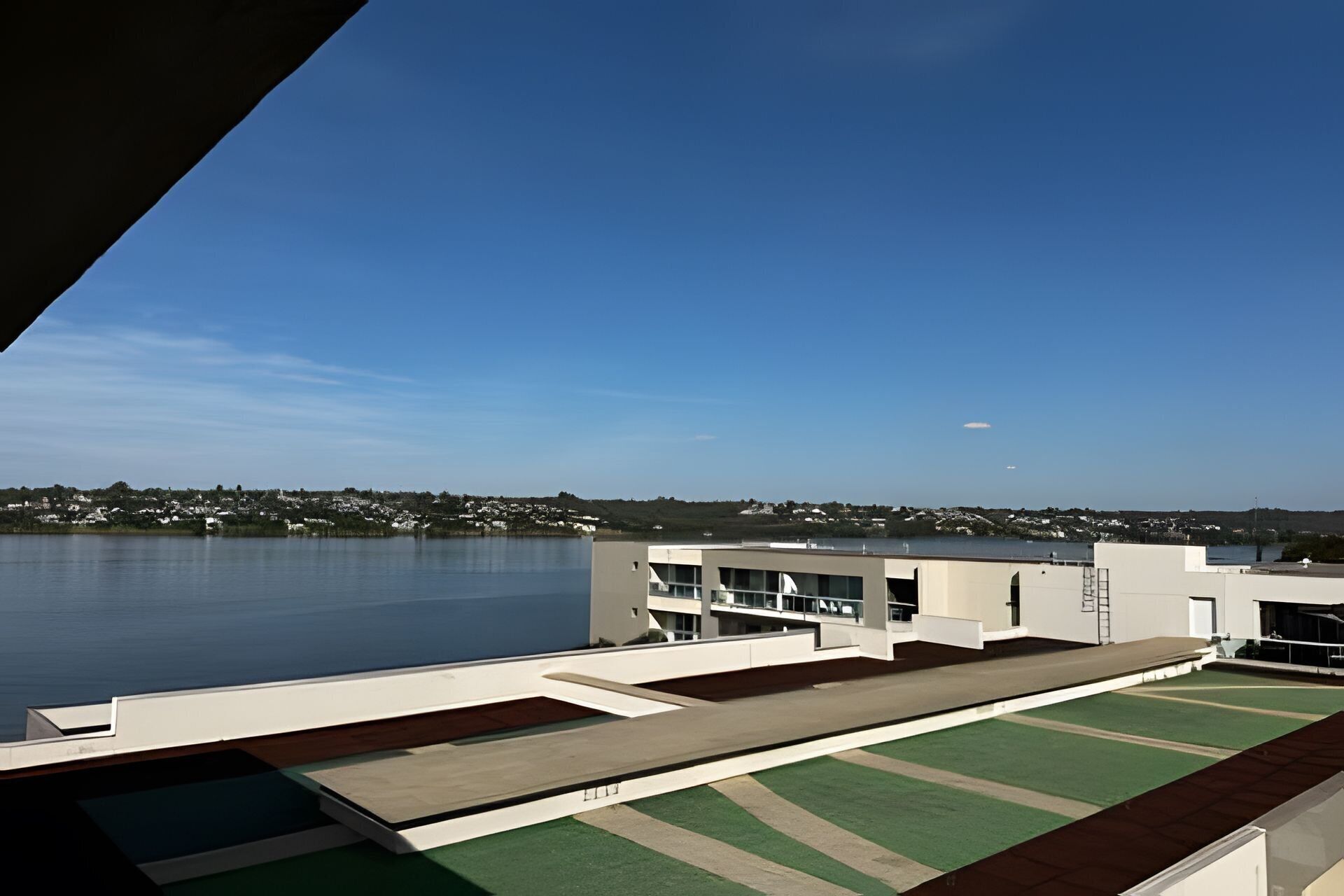 A modern building with a flat roof overlooking a calm body of water and a distant shoreline.