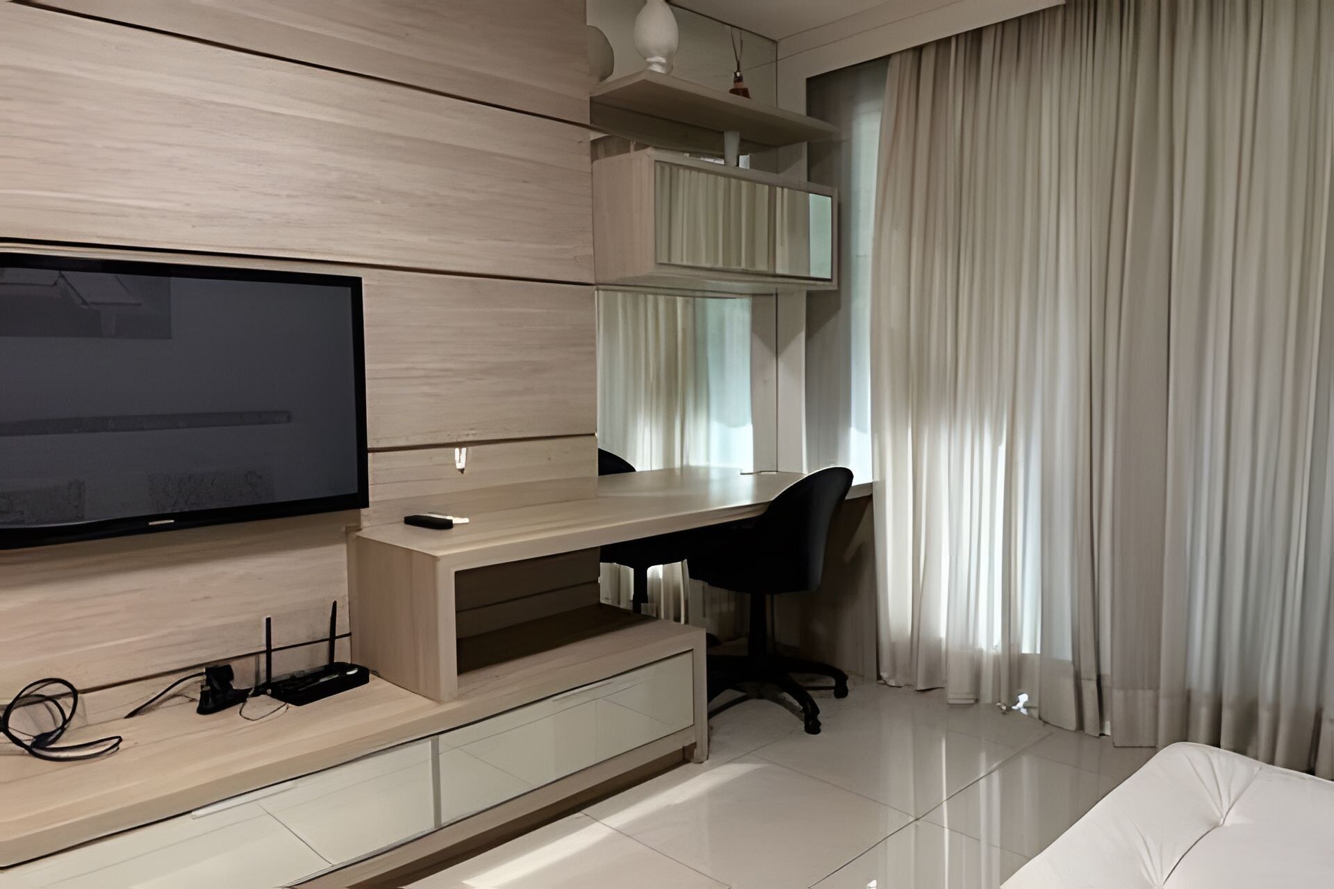 A modern living room with a flat-screen TV mounted on a wooden wall, a desk with a chair, and a large window with sheer curtains.