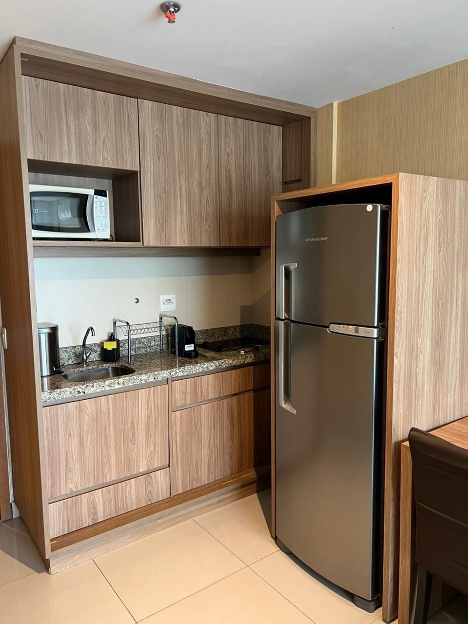 A compact kitchen with wooden cabinets, a stainless steel refrigerator, a microwave, and a sink with modern fixtures.