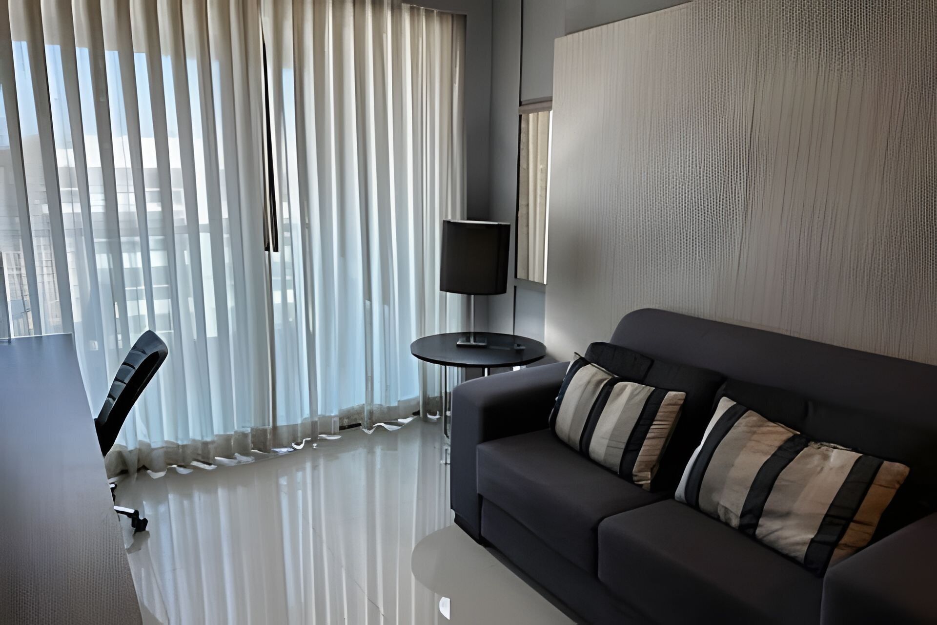 Uma sala de estar moderna com um sofá escuro, almofadas listradas, uma pequena mesa redonda com uma luminária e cortinas leves.