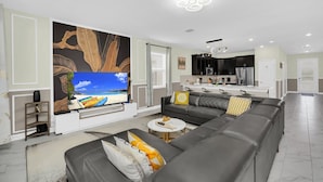 Een moderne woonkamer met een grote flatscreen televisie, een grijze modulaire bank en een eethoek, met een keuken op de achtergrond.