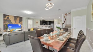Een moderne eetruimte met een houten tafel gedekt voor zes personen, omringd door lederen stoelen. In de achtergrond is een keuken met donkere kasten en een magnetron zichtbaar.