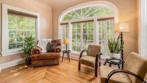 A cozy room with a brown leather sofa, a beige armchair, and a small table with a lamp. There are two windows with views of greenery outside. - 3/2 Luxury Spanish Villa - Pool - Pet Friendly (Lakeland)