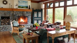 Un salon chaleureux avec une cheminée en pierre, des sièges confortables et de grandes fenêtres donnant sur l’extérieur.