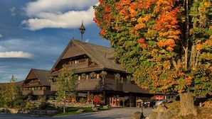 Un chalet traditionnel en bois, avec un bâtiment central imposant et deux annexes de chaque côté, entouré d’un feuillage automnal et sous un ciel dégagé.