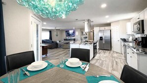 Una cocina moderna con refrigerador empotrado, un comedor con mesa lista para dos comensales y una sala de estar con sofá y televisor.