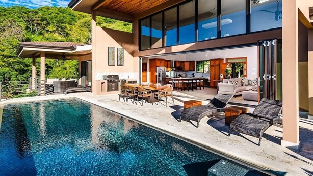 Una casa moderna con piscina, zona de comedor exterior y una cocina amplia visible a través de puertas de cristal.