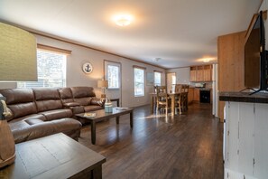 A spacious living room with a leather sofa, wooden coffee table, and a dining area with a wooden table and chairs. A kitchen with wooden cabinets and a refrigerator is visible in the background.