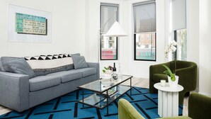Una sala moderna con un sofá gris, una mesa de centro de cristal y un sillón verde. Hay un boleto enmarcado en la pared y una ventana con vistas a edificios.