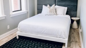 Una cama bien hecha con ropa de cama blanca y dos almohadas decorativas, una mesita de noche y una ventana con vista a un muro de ladrillo.