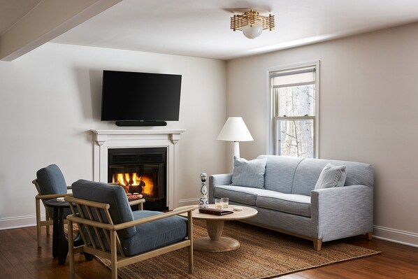 A living room with a fireplace, a television mounted above it, a sofa, a chair, and a lamp. - The Neighborhood Hotel Grand Beach Suite 12 - Three Bedroom (Grand Beach)
