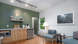 Et moderne køkken med træskabe, indbygget mikrobølgeovn og et rustfrit stål køleskab. Der er et spiseområde med en blå sofa og et lille bord. Et vindue giver udsigt til grønne omgivelser udenfor.