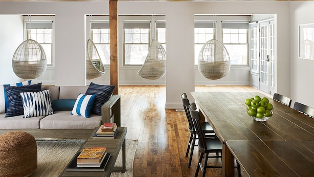 A modern living room with a sofa, coffee table, and hanging wicker chairs. There is a dining table with chairs and a bowl of green apples.