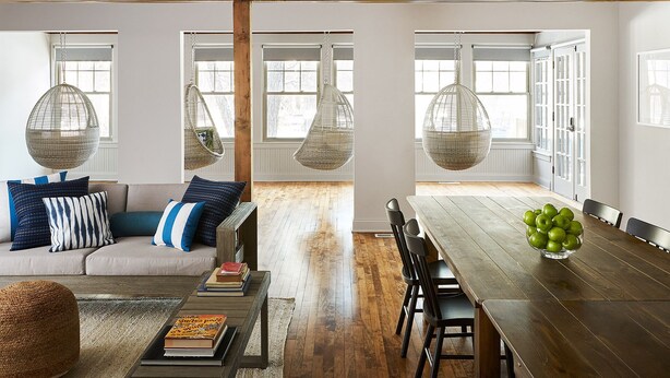 A modern living room with a sofa, coffee table, and hanging wicker chairs. There is a dining table with chairs and a bowl of green apples.