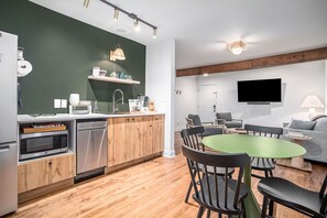 Une cuisine moderne avec des meubles en bois, un coin repas doté d’une table verte et un salon avec un canapé gris et un téléviseur à écran plat.