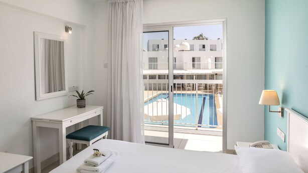 Ein Hotelzimmer mit Bett, Schreibtisch, Stuhl, Nachttisch, Lampe, Telefon und einem Fenster mit Blick auf einen Pool und Gebäude.