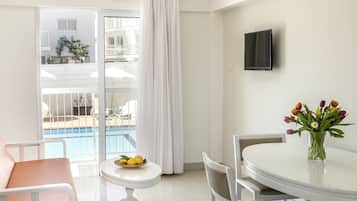 Ein modernes Hotelzimmer mit Balkon, einem Essbereich inklusive Tisch und Stühlen, einem kleinen runden Tisch mit Obstschale und einer Vase mit Blumen.