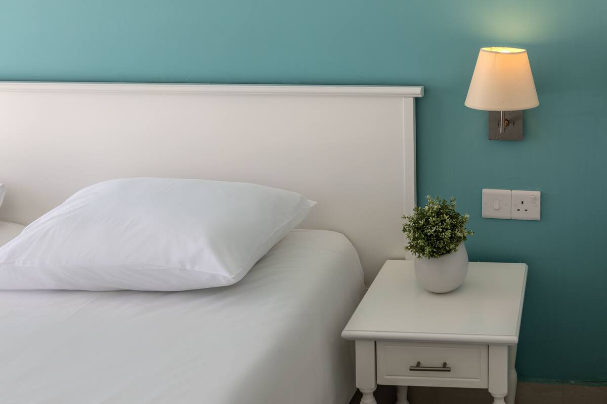 Ein ordentlich bezogenes Bett mit weißer Bettwäsche und Kissen, ein kleiner Nachttisch mit Topfpflanze und eine Wandlampe.