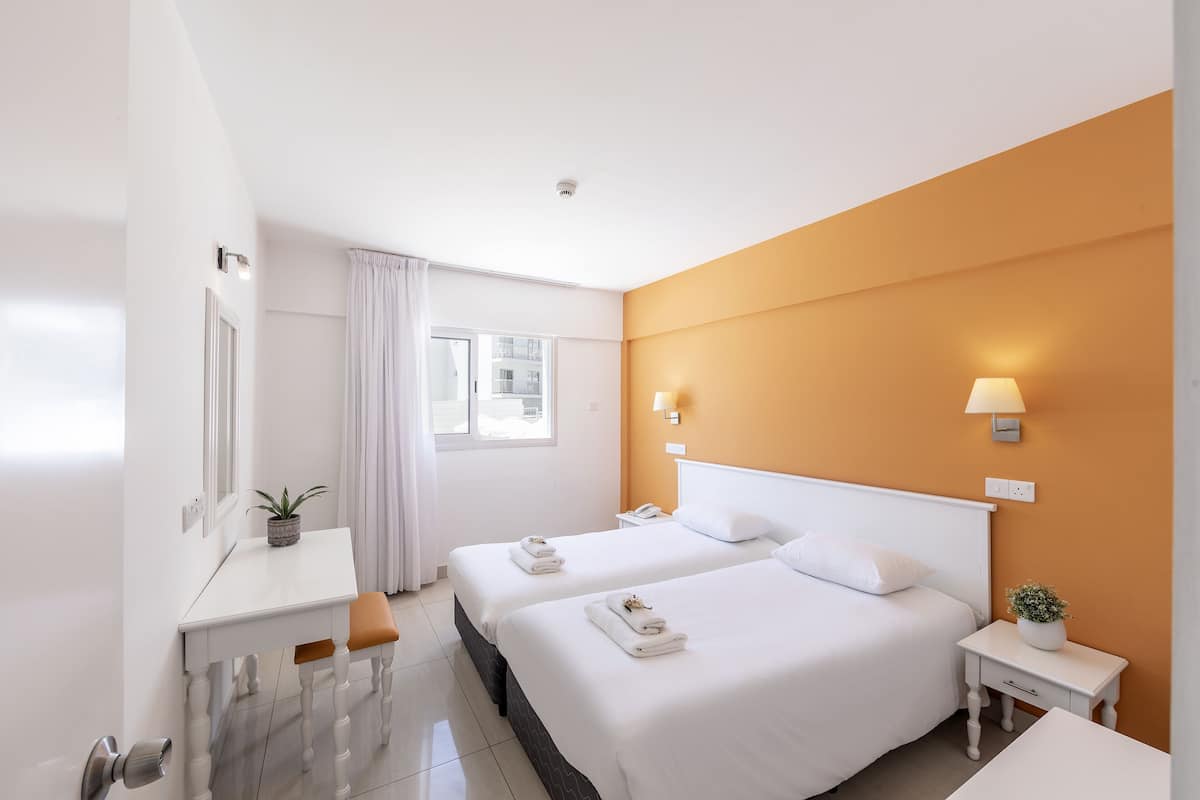 Ein Hotelzimmer mit zwei Betten, einem Schreibtisch, einem Stuhl, einem Nachttisch, einer Lampe und einem Fenster.