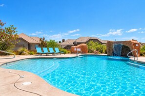 Una piscina con caída de agua, rodeada de sillones de descanso y un cielo despejado.
