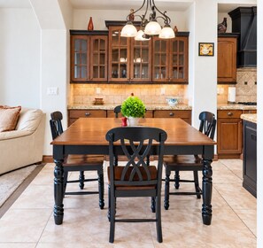 Een eetruimte met een houten tafel en stoelen, een plant in pot en een keuken met houten kasten en een kroonluchter.