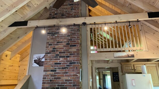 A rustic interior with exposed wooden beams, a brick fireplace, and a ceiling fan. - Log cabin~hike, hunt, ski,with direct snowmobile trail access and outdoor sauna. (Upton)