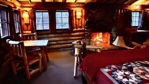 Ein gemütliches Blockhaus-Interieur mit Holzbalkendecke, Kamin, Essbereich und einem Bett mit Steppdecke.