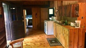 Een blokhutinterieur met een houten vloer, een kitchenette met fornuis en koelkast, en een deur die leidt naar een slaapkamer.