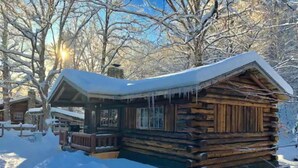 Uma cabana de madeira com telhado coberto de neve e varanda, rodeada por árvores e neve.