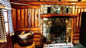 Interior de uma cabana de troncos com lareira de pedra, paredes de madeira e cortina xadrez.