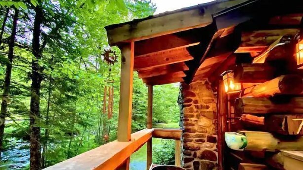 Un chalet rustique doté d’une terrasse en bois et de murs en pierre, entouré d’une végétation luxuriante.