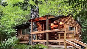 Une cabane rustique en rondins de bois, dotée d’une cheminée en pierre, d’une terrasse en bois et d’un escalier menant à l’entrée.