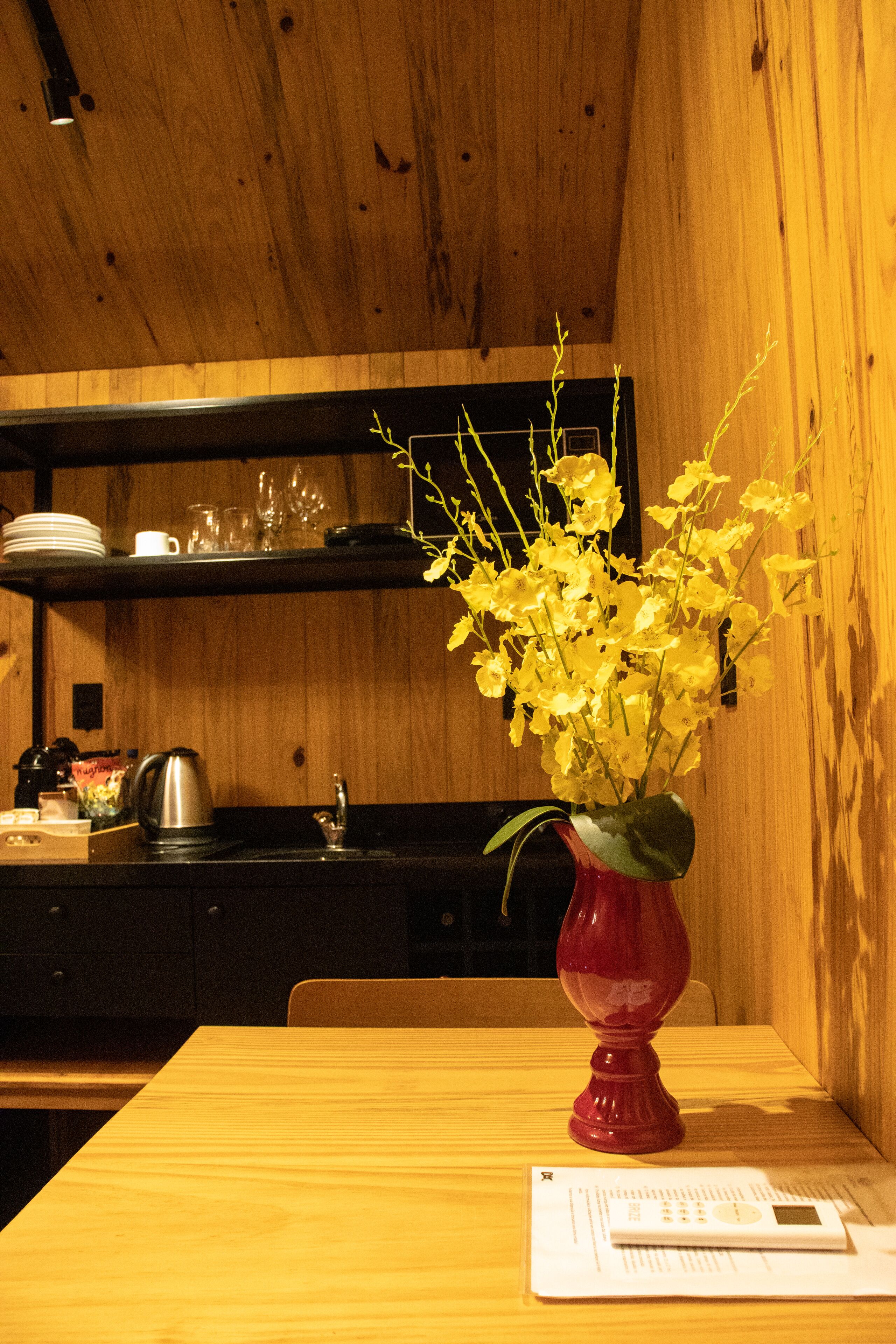Un interior de madera con una mesa, un jarrón con flores amarillas y utensilios de cocina en estantes.